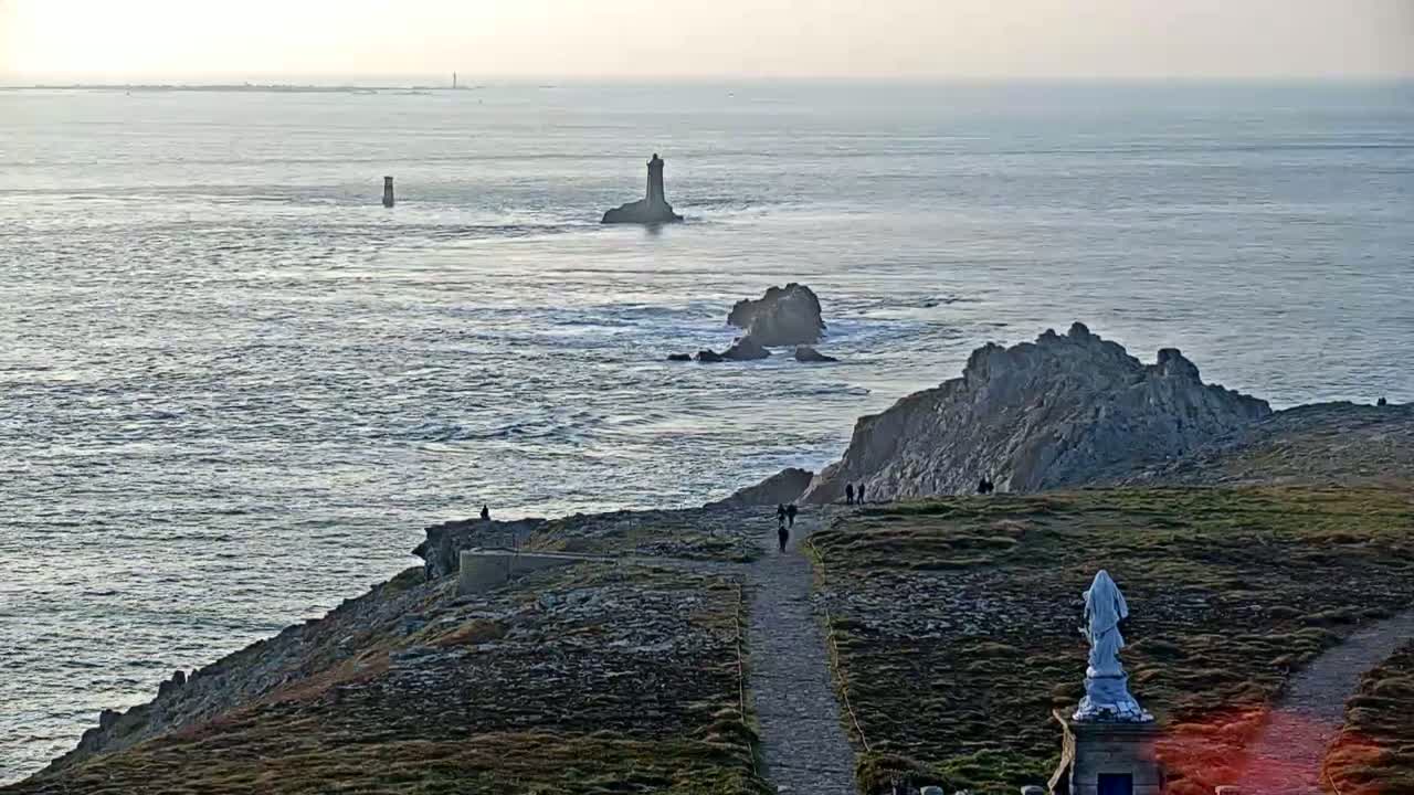 A coastal path winds through a rocky landscape overlooking a choppy sea with several small islands and lighthouses under a hazy sky.