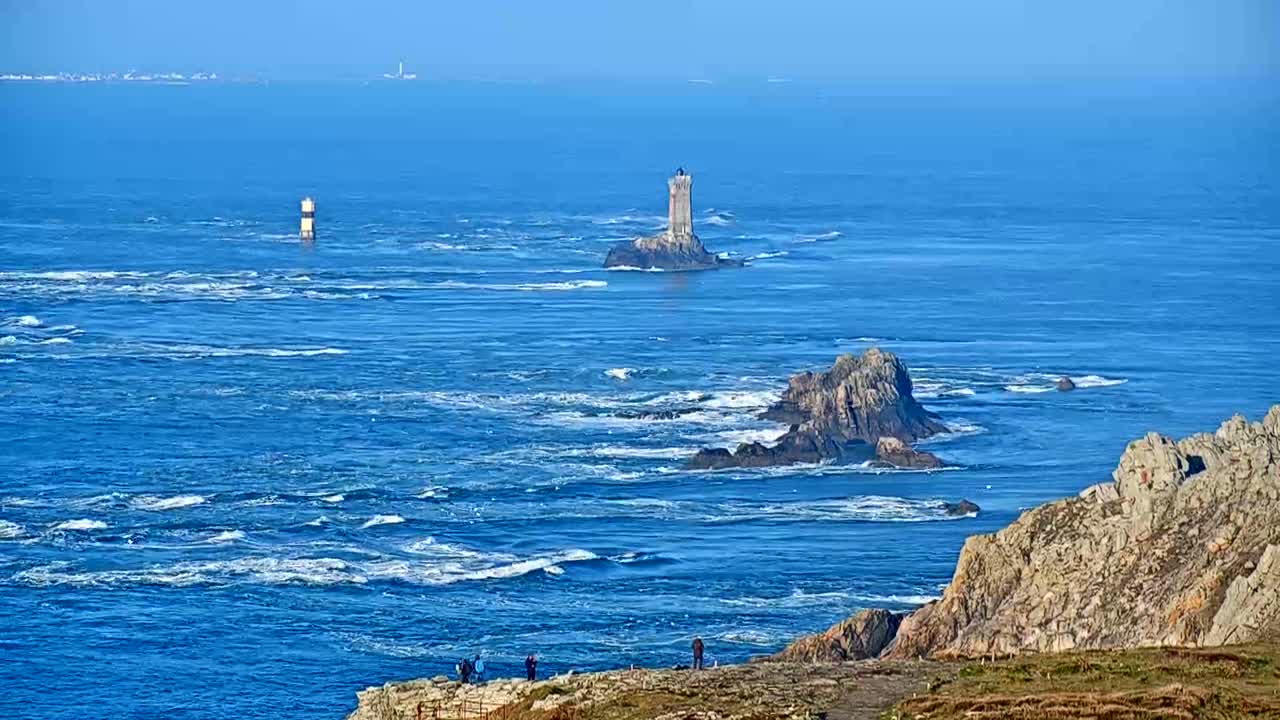 A rocky coastline with a lighthouse on a small island in the distance, under a clear blue sky with a moderately choppy sea.