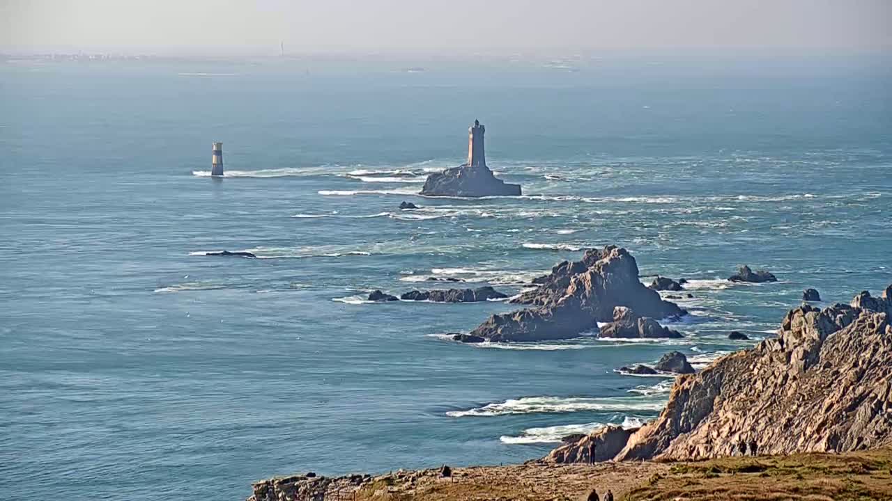 A distant lighthouse and rocky coastline are visible on a hazy, sunny day with a choppy sea.
