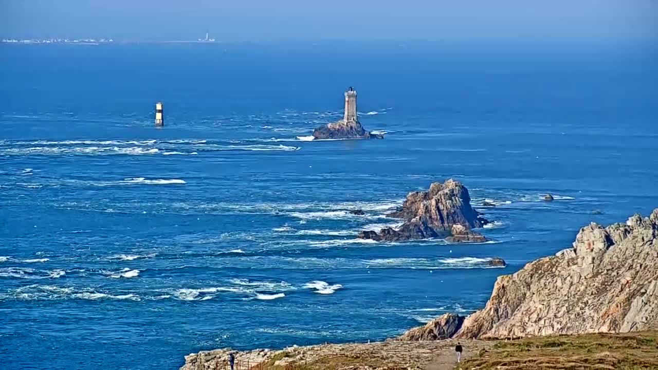 A rocky coastline with two lighthouses visible in the distance, under a clear blue sky, with a choppy ocean.