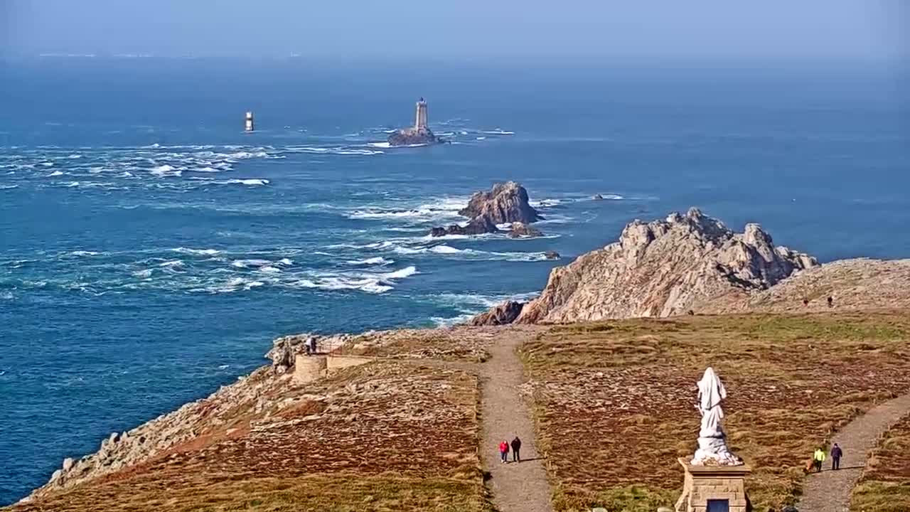 A coastal overlook on a sunny day shows a path leading to a statue, with the ocean displaying whitecaps and two lighthouses visible in the distance.