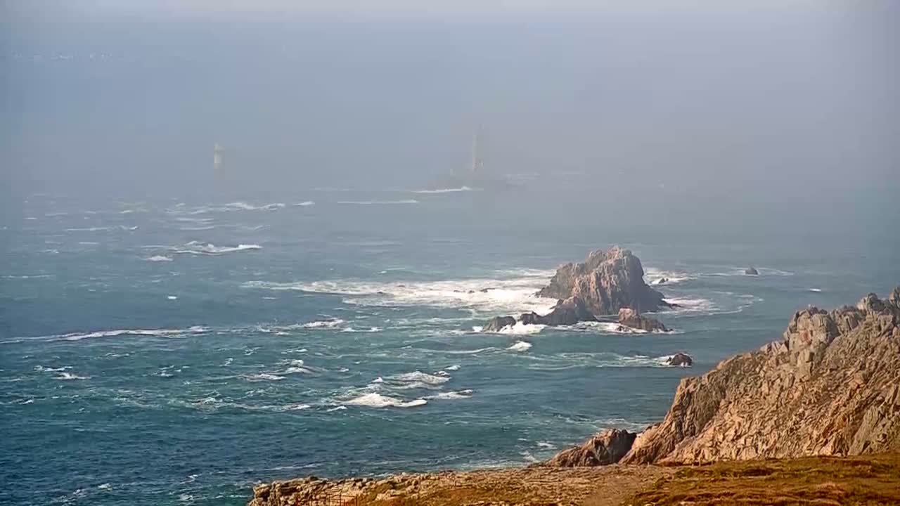 A hazy, somewhat choppy ocean with rocky outcroppings and distant lighthouses is visible from a coastal cliff.