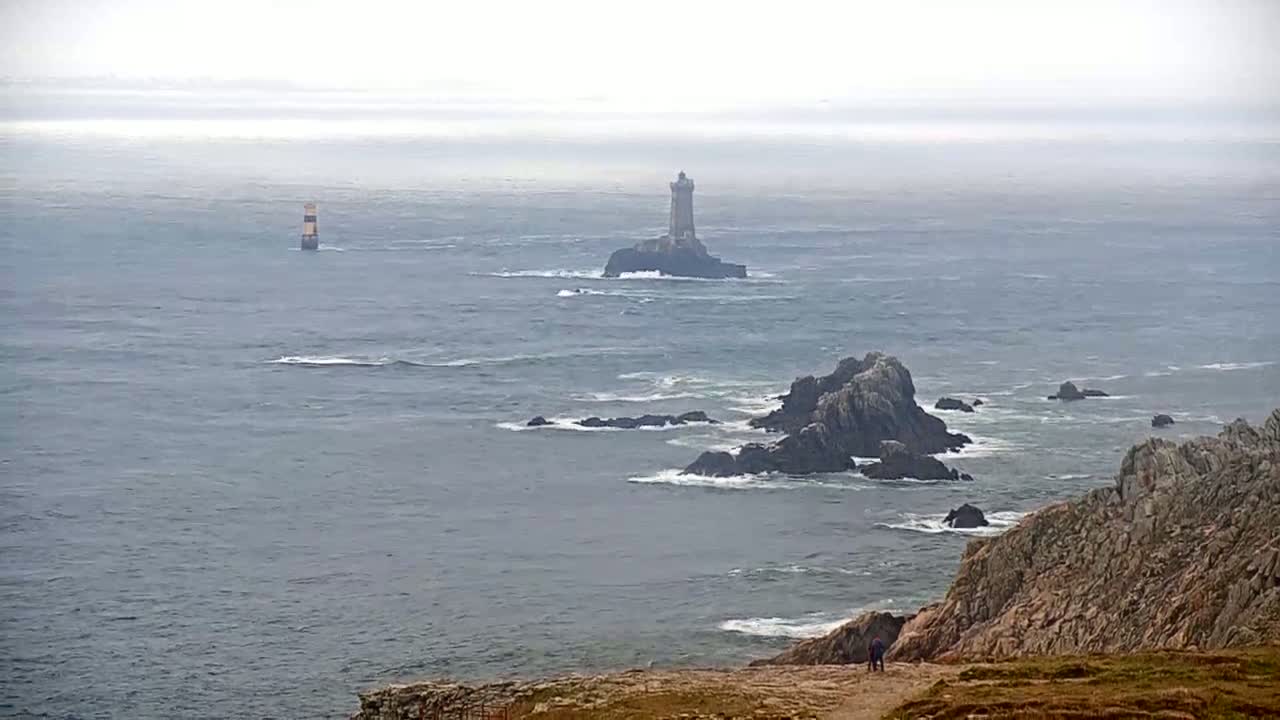 A foggy coastal scene shows a lighthouse on a small island in the distance, with rocky outcrops and a person standing on a cliff in the foreground.
