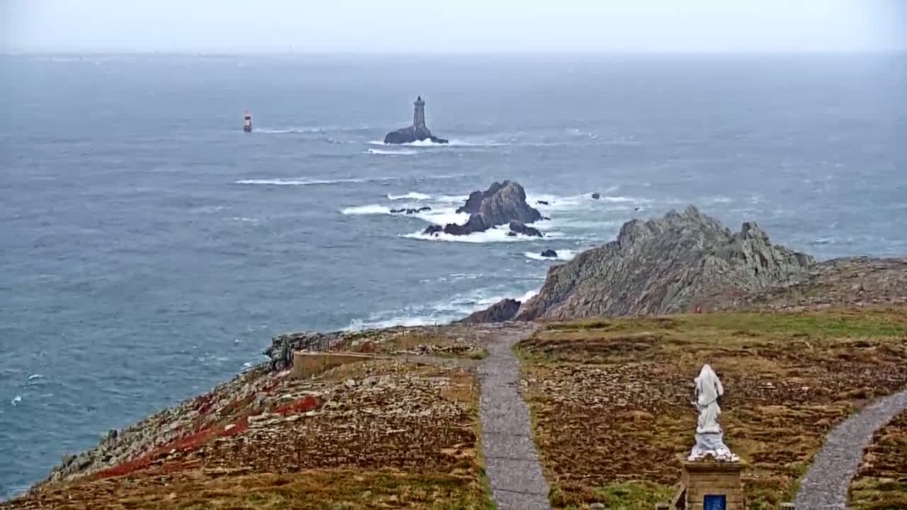 A rocky coastline with a lighthouse in the distance, a statue on a path overlooking the churning sea under an overcast sky.