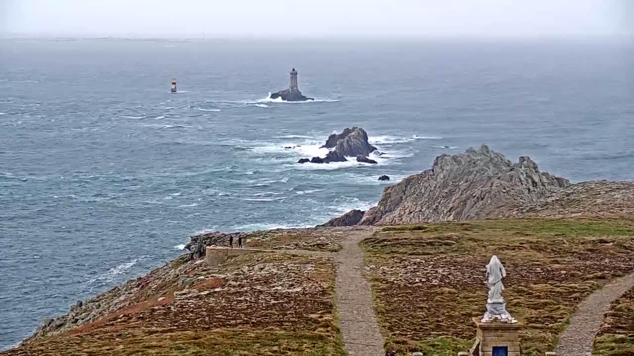 A rocky coastal scene on an overcast day shows a statue, paths, and a distant lighthouse on a small island, with choppy waves visible in the ocean.