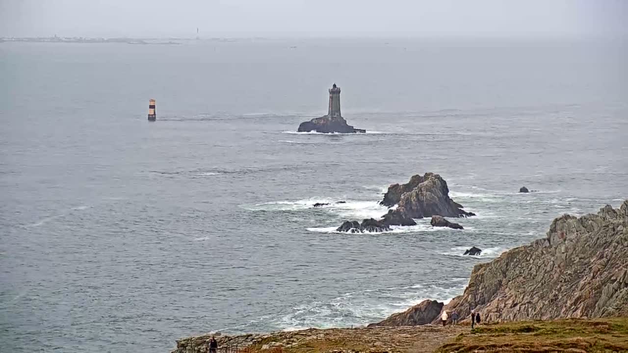 A lighthouse sits on a small island in a gray, overcast ocean, with rocky outcrops and a few people visible on a nearby cliff.