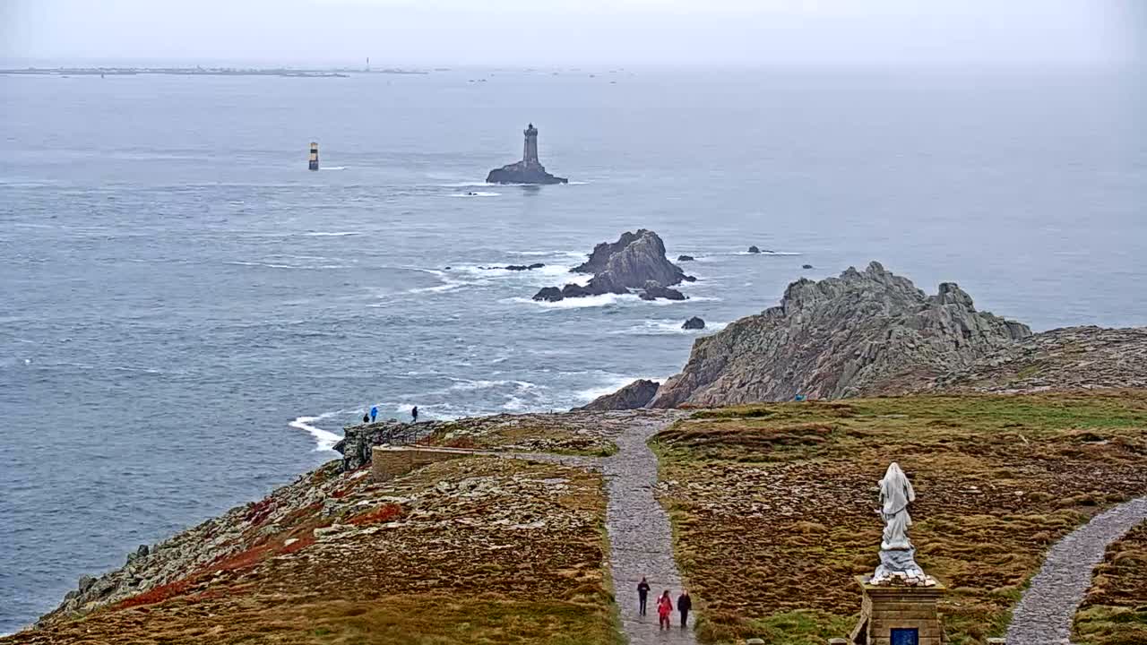 A coastal path winds down a rocky outcrop overlooking a choppy sea dotted with islands and lighthouses, under an overcast sky.