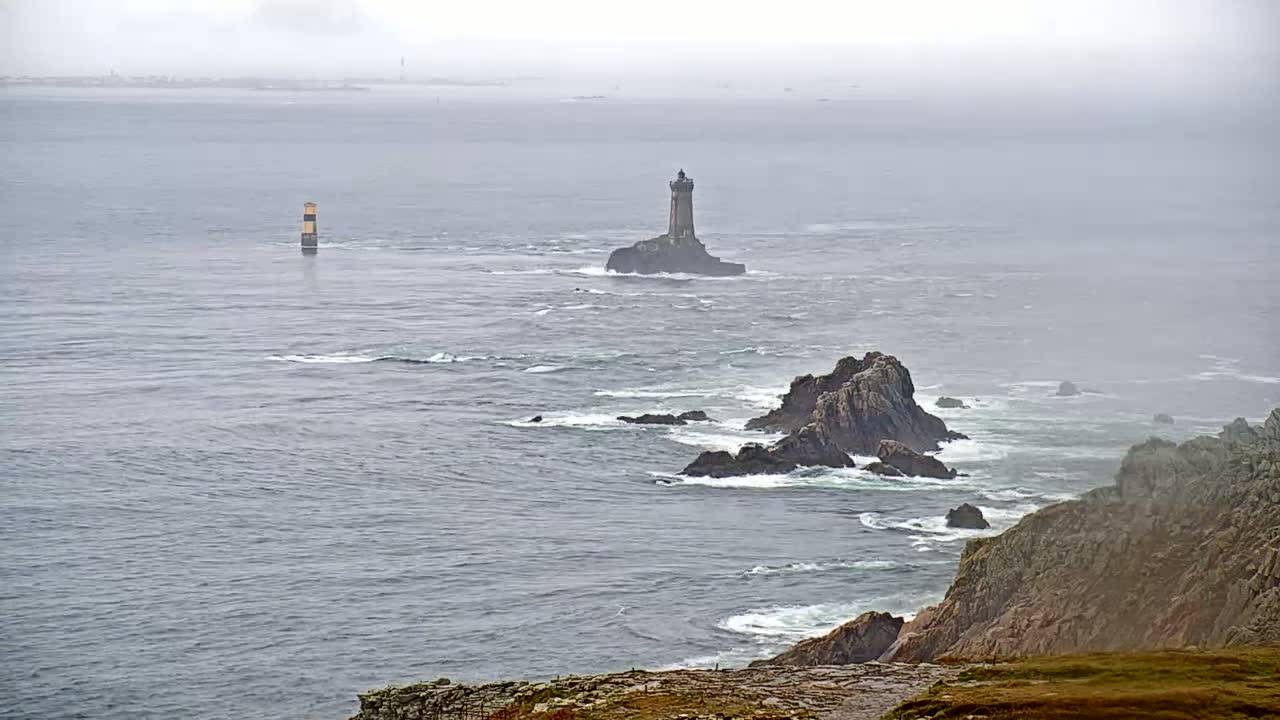 A lighthouse sits on a small island in a foggy, grey sea, with rocky coastlines visible in the foreground and background.