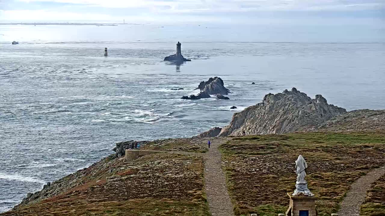A rocky coastline with a statue overlooking a choppy sea, featuring distant islands and lighthouses under a cloudy sky.