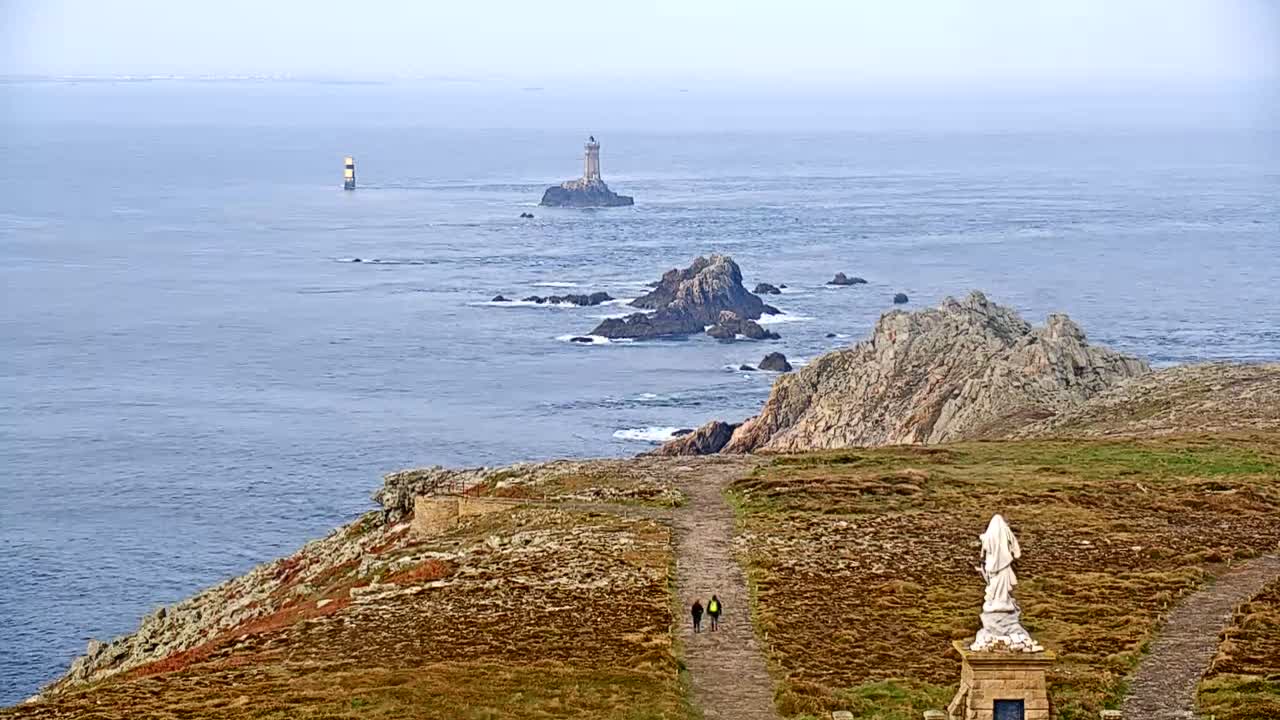 A coastal path leads to a statue overlooking a rocky coastline and distant lighthouses on a hazy day.