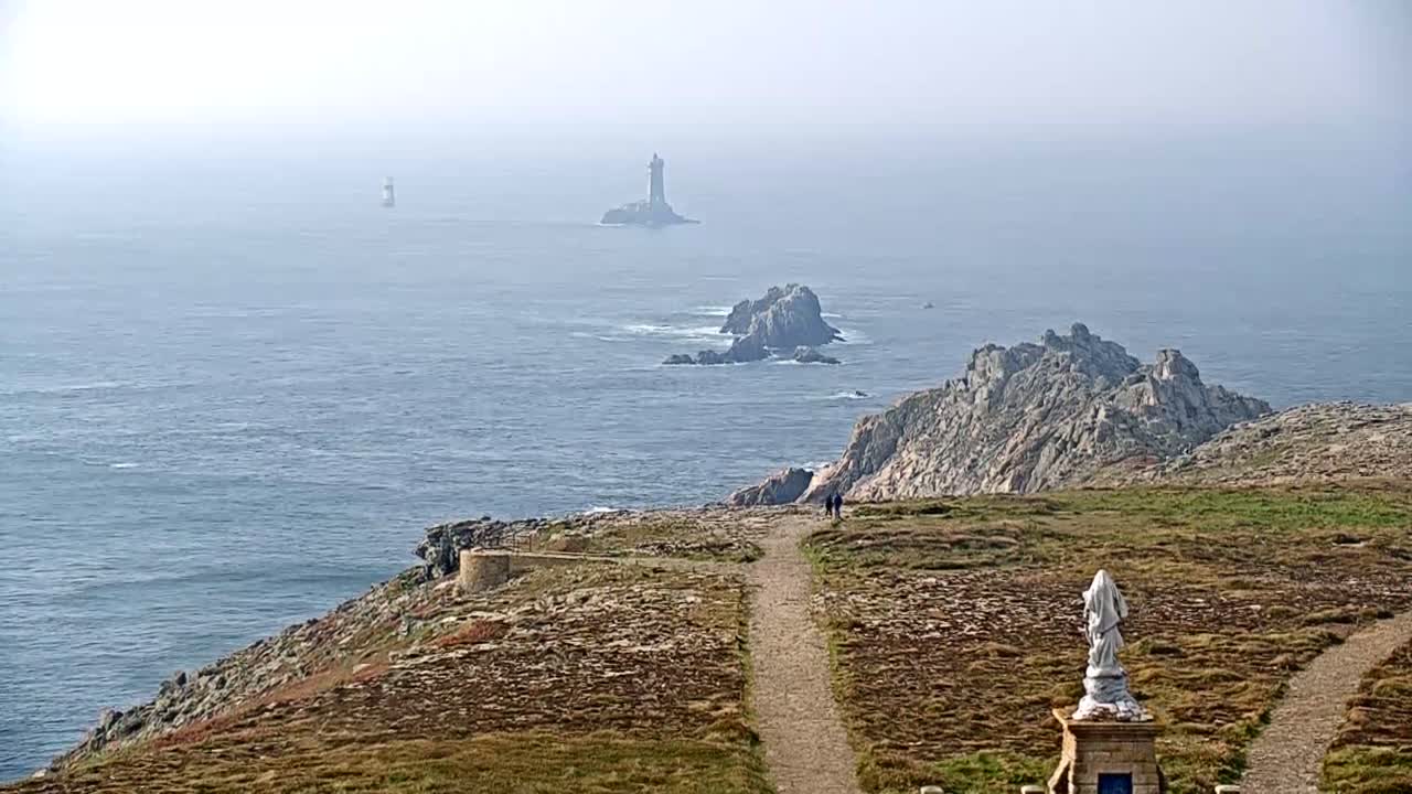 A hazy coastal scene shows a rocky, grassy cliff overlooking the ocean with two lighthouses and several rocky islands visible in the distance.