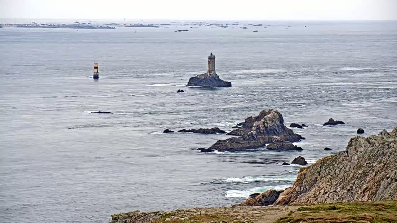 A stone lighthouse sits on a small island in a choppy sea, with rocky coastlines and other navigational markers visible in the distance under an overcast sky.