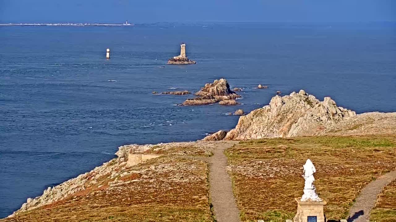 A coastal scene on a clear day shows a rocky shoreline with a statue, a path, and distant islands with lighthouses in the calm ocean.