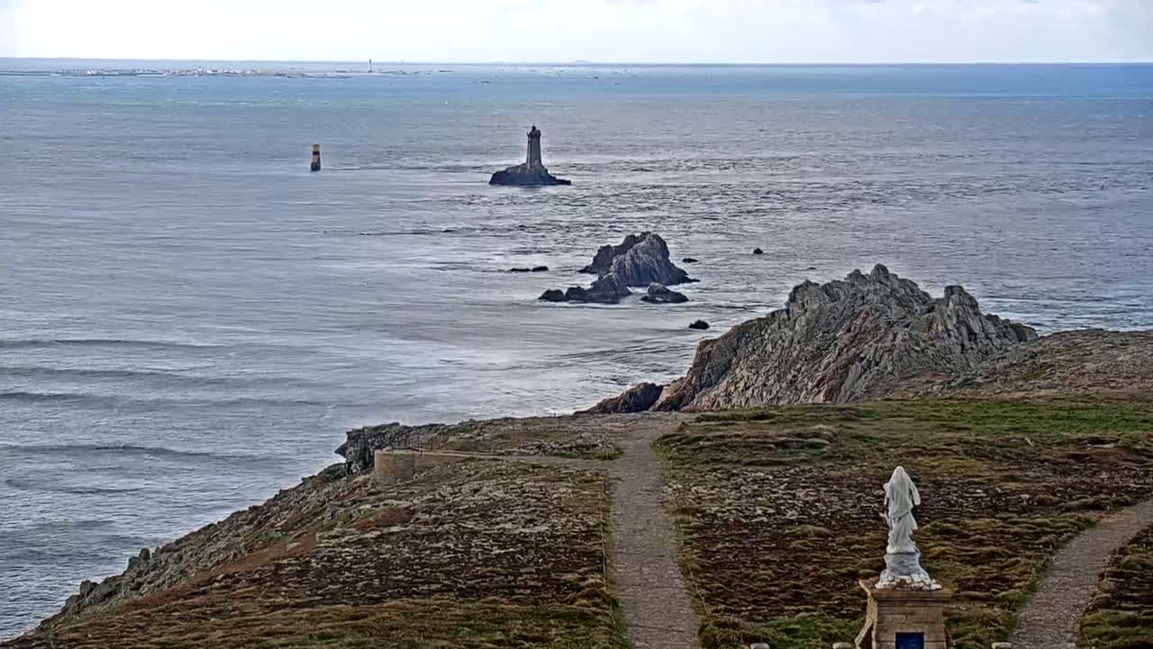 A coastal scene with a statue overlooking a rocky shoreline and ocean with small waves, featuring a distant lighthouse and a smaller marker in the water under a mostly cloudy sky.