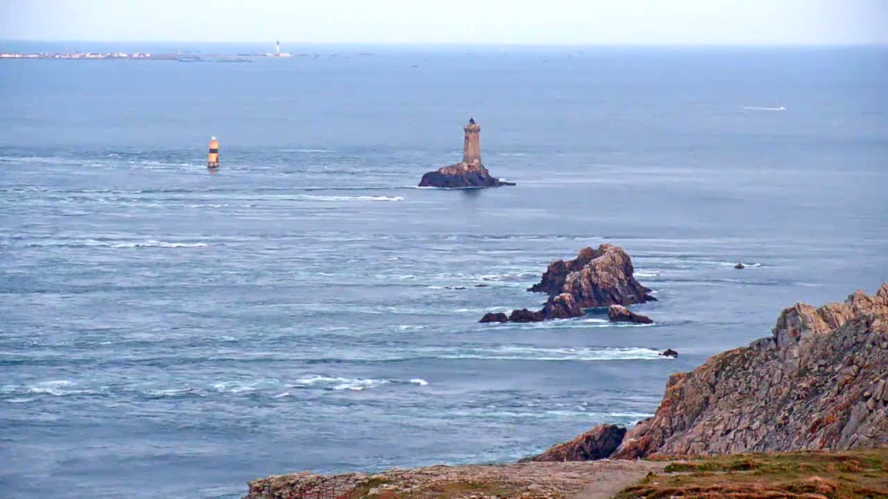 A rocky coastline overlooks a turbulent sea with two lighthouses and small islands visible in the distance under a hazy sky.