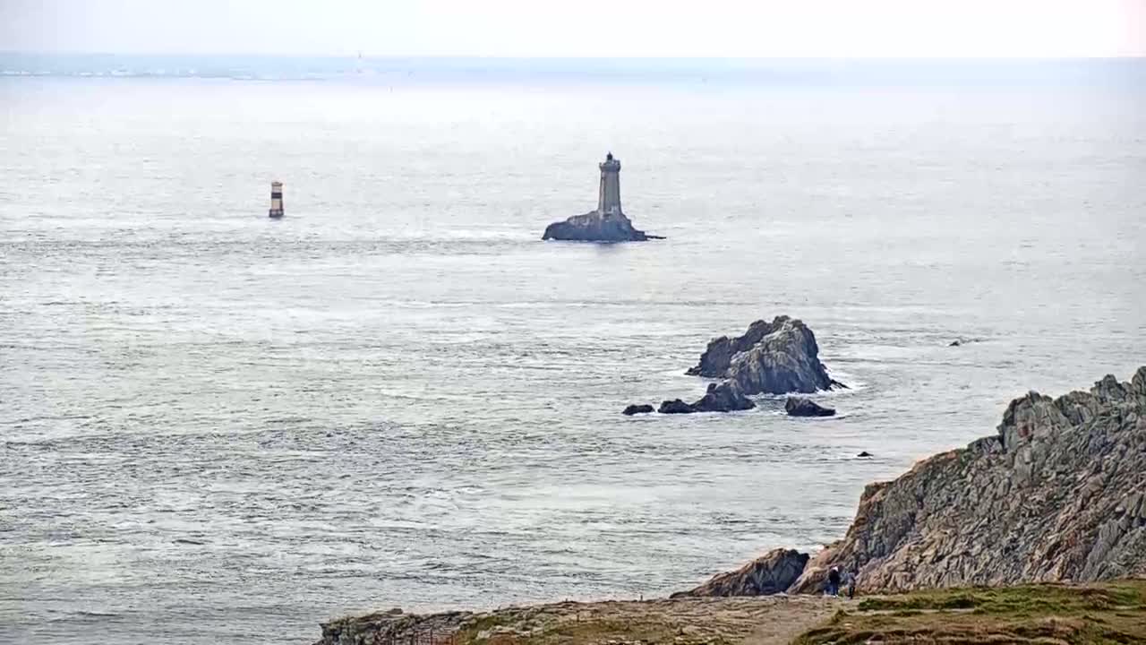 A rocky coast under an overcast sky features a lighthouse on a small island and another, smaller marker further out to sea.