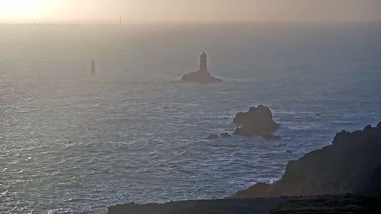 A hazy sunset illuminates a rocky coastline with two lighthouses and smaller rock formations visible in the distance across a choppy sea.
