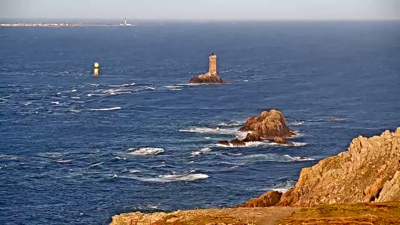 A rocky coastline with two lighthouses visible in the distance, under a mostly clear sky with a somewhat choppy sea.