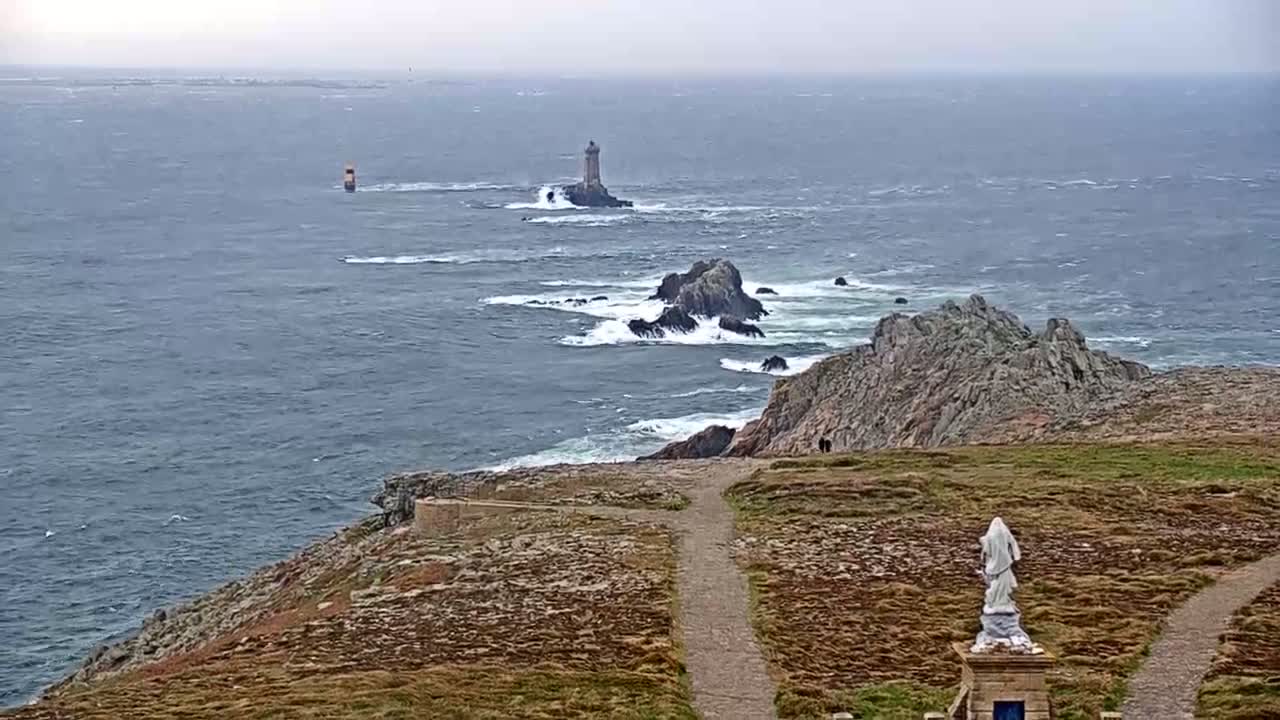 A statue stands on a grassy cliff overlooking a choppy sea with distant lighthouses and rocky outcrops under an overcast sky.