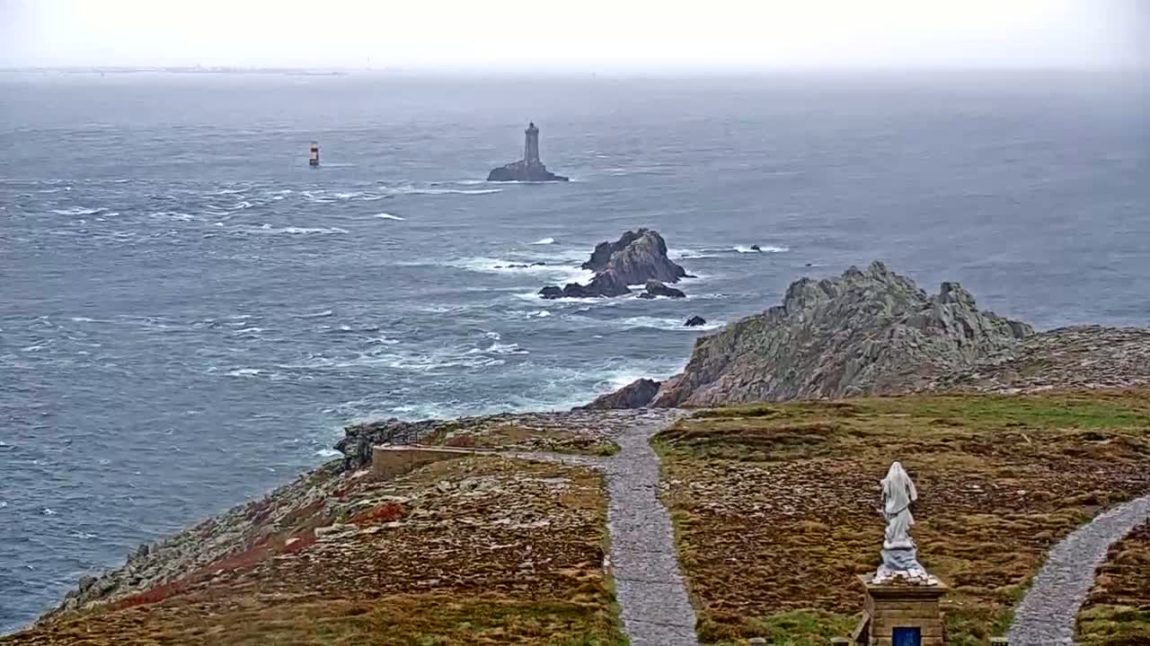 A statue stands on a rocky, grassy path overlooking a choppy ocean with distant islands and a lighthouse under an overcast sky.