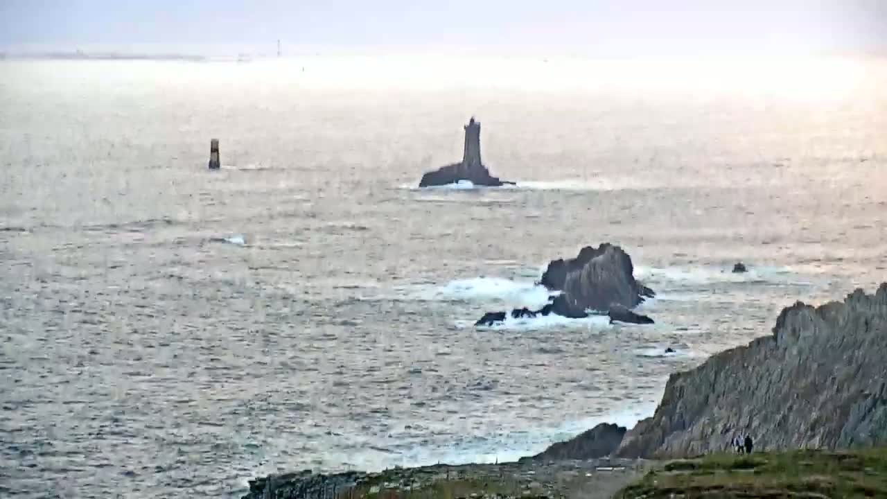 A rocky coastline with several small islands and a tower-like structure visible in the distance under an overcast sky, with waves breaking against the rocks.