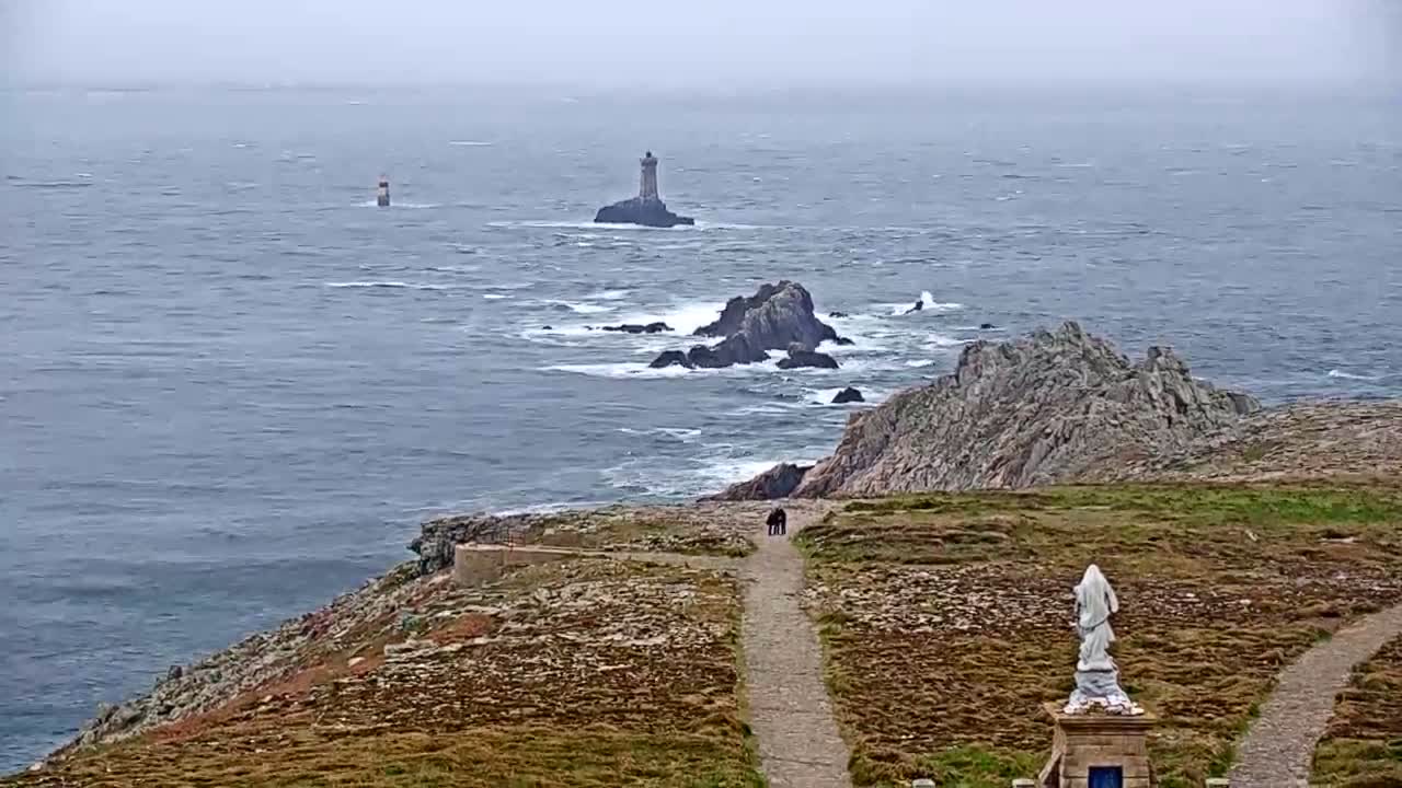 A coastal scene on an overcast day shows a rocky shoreline with two pathways leading to a statue, overlooking a choppy ocean with two lighthouses in the distance.