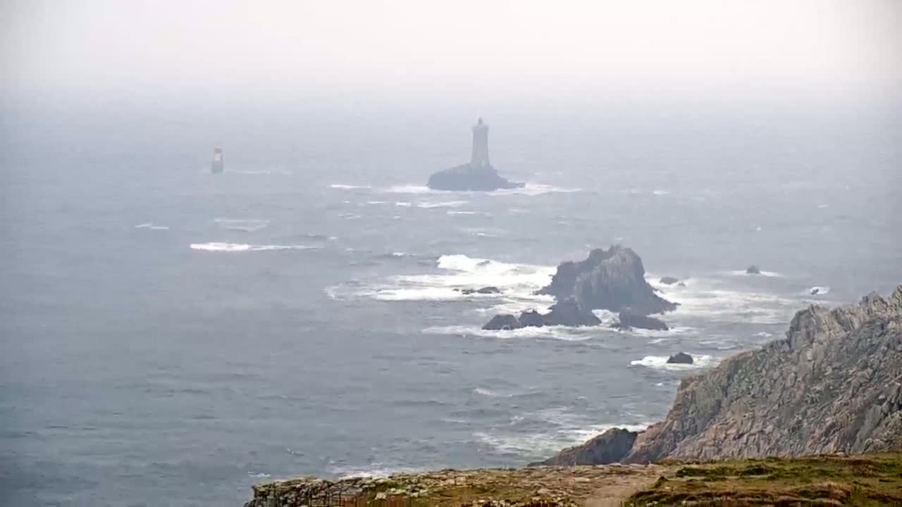 A distant lighthouse is visible on a rocky outcrop in a foggy, wavy ocean, with closer rocky cliffs and waves in the foreground.