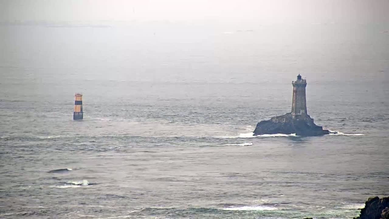 A tall lighthouse sits on a small, rocky island in a foggy ocean, with a smaller, striped marker farther away.
