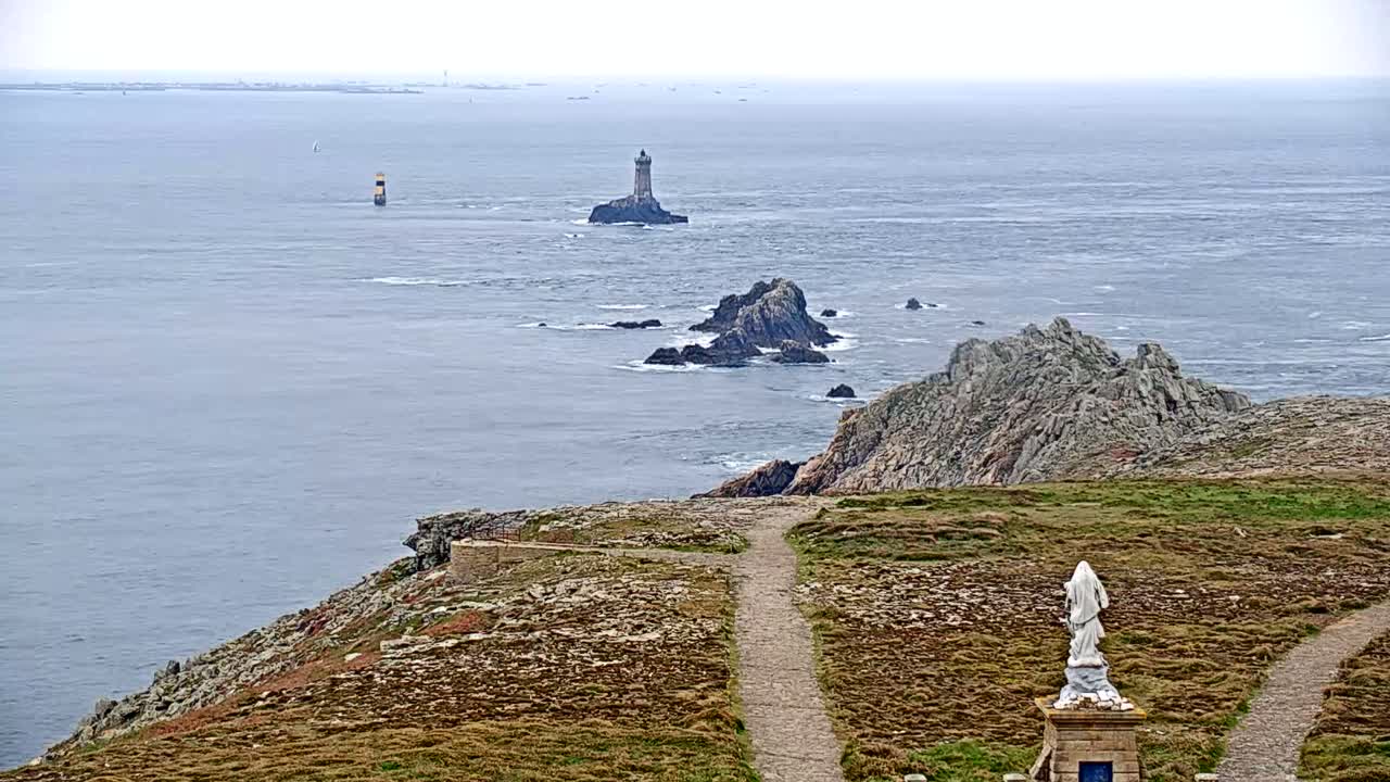 A statue overlooks a coastal path and rocky outcroppings, with a lighthouse visible in the distance across a calm, gray sea under an overcast sky.
