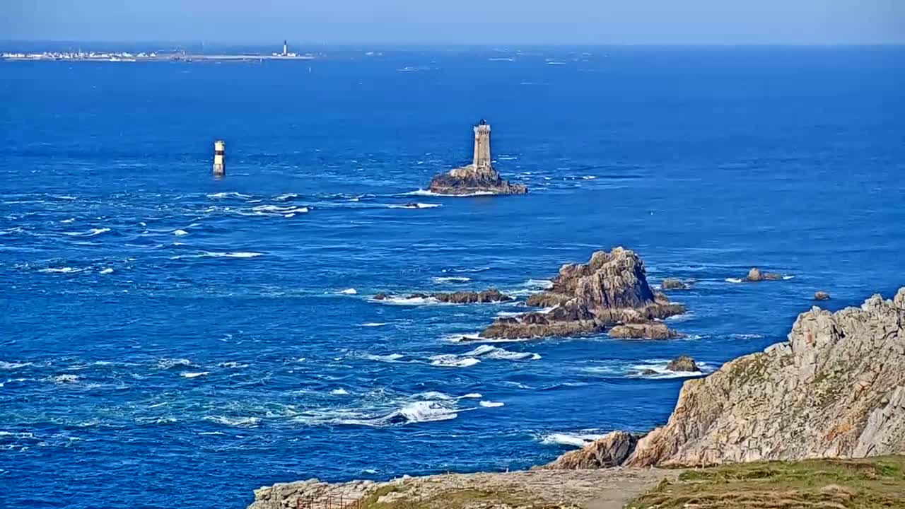 A rocky coastline with two lighthouses visible on the ocean under sunny conditions, with choppy waves breaking against the rocks.