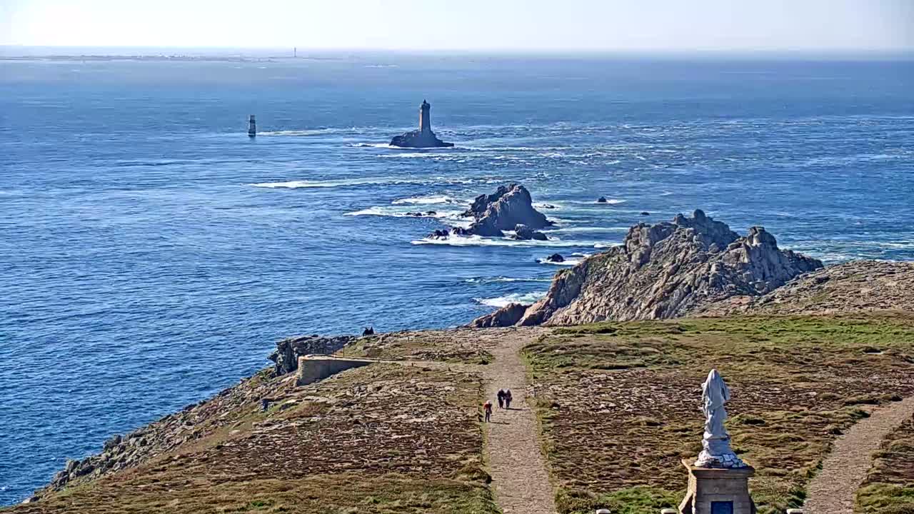 A coastal path winds through rocky, grassy terrain overlooking a blue ocean dotted with islands, one featuring a lighthouse, under sunny skies.