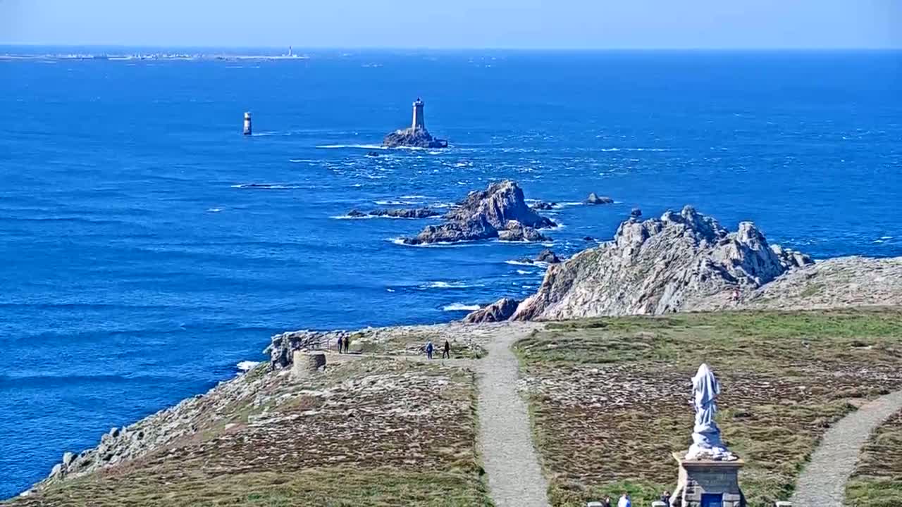 A rocky coastline with a statue and a path overlooks a blue ocean dotted with small islands and distant lighthouses under sunny skies.