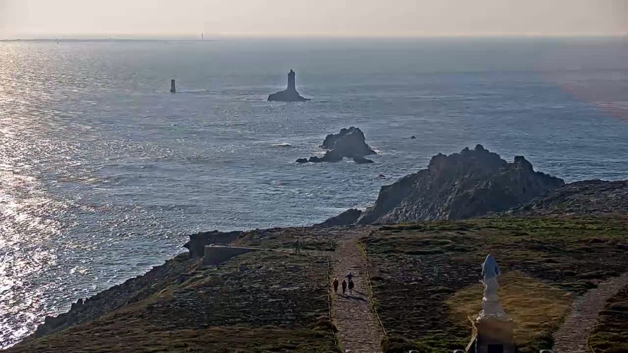 A coastal scene on a partly sunny day shows a rocky shoreline with a paved path leading to a statue, and distant islands with lighthouses visible across a calm sea.