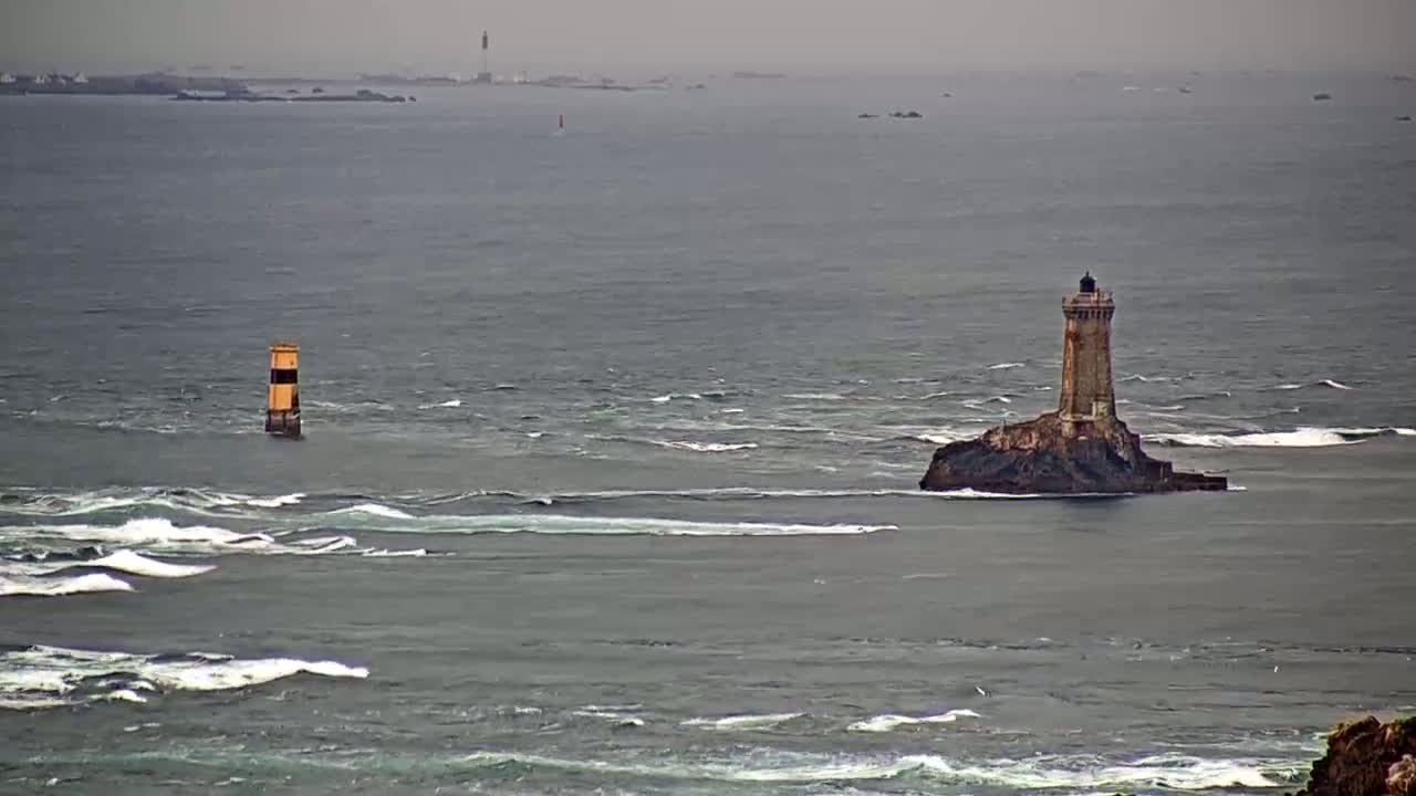 A stone lighthouse sits on a small, rocky island in a choppy sea under an overcast sky.