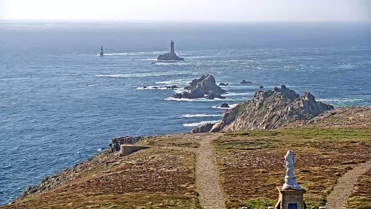 A coastal vista shows a rocky outcrop with a lighthouse in the distance, under a hazy, sunny sky, and a statue near the foreground.