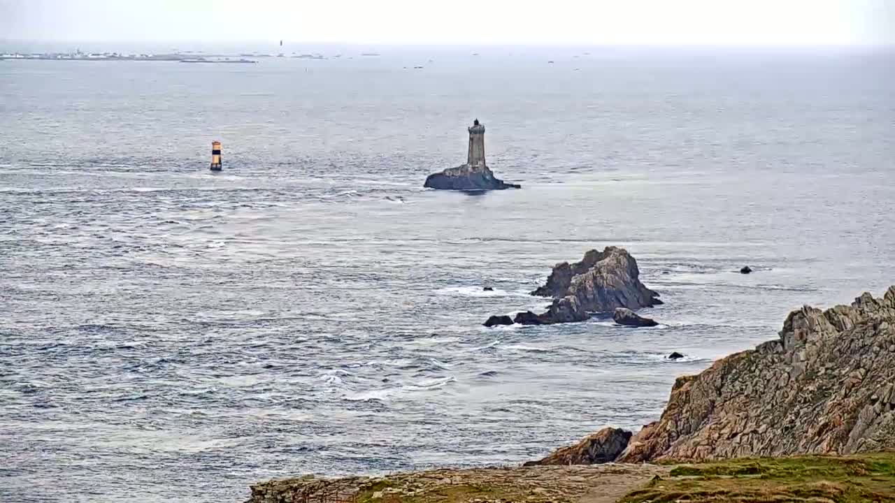 A coastal scene on an overcast day shows a lighthouse on a small island in the distance, with rocky outcrops and choppy water closer to the foreground.