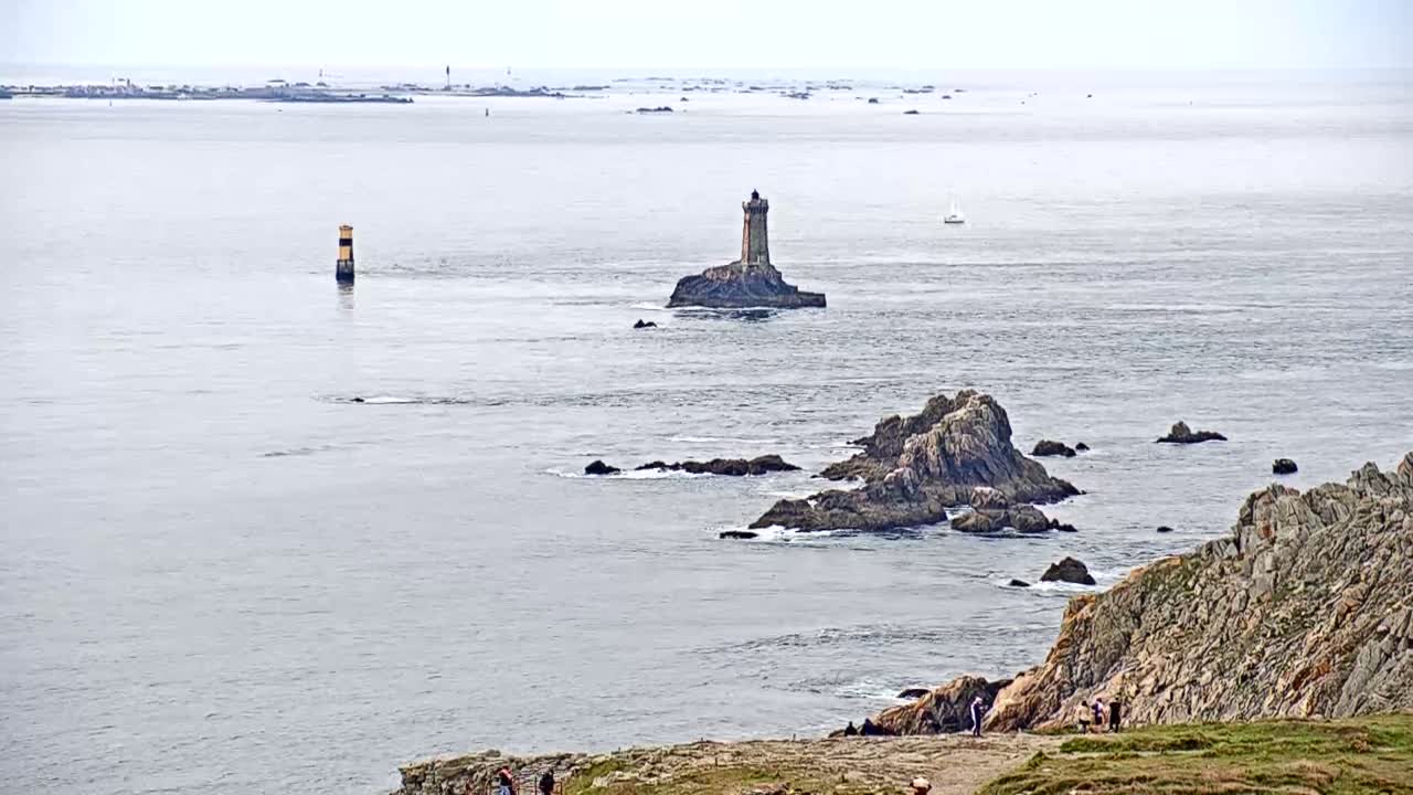 A lighthouse sits on a small island in a calm, gray sea, surrounded by rocky outcrops and a distant coastline under an overcast sky.