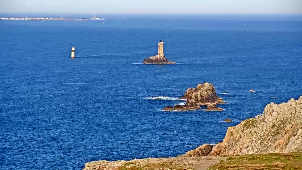 A rocky coastline with two lighthouses and several small islands is visible on a clear, sunny day over a deep blue ocean.