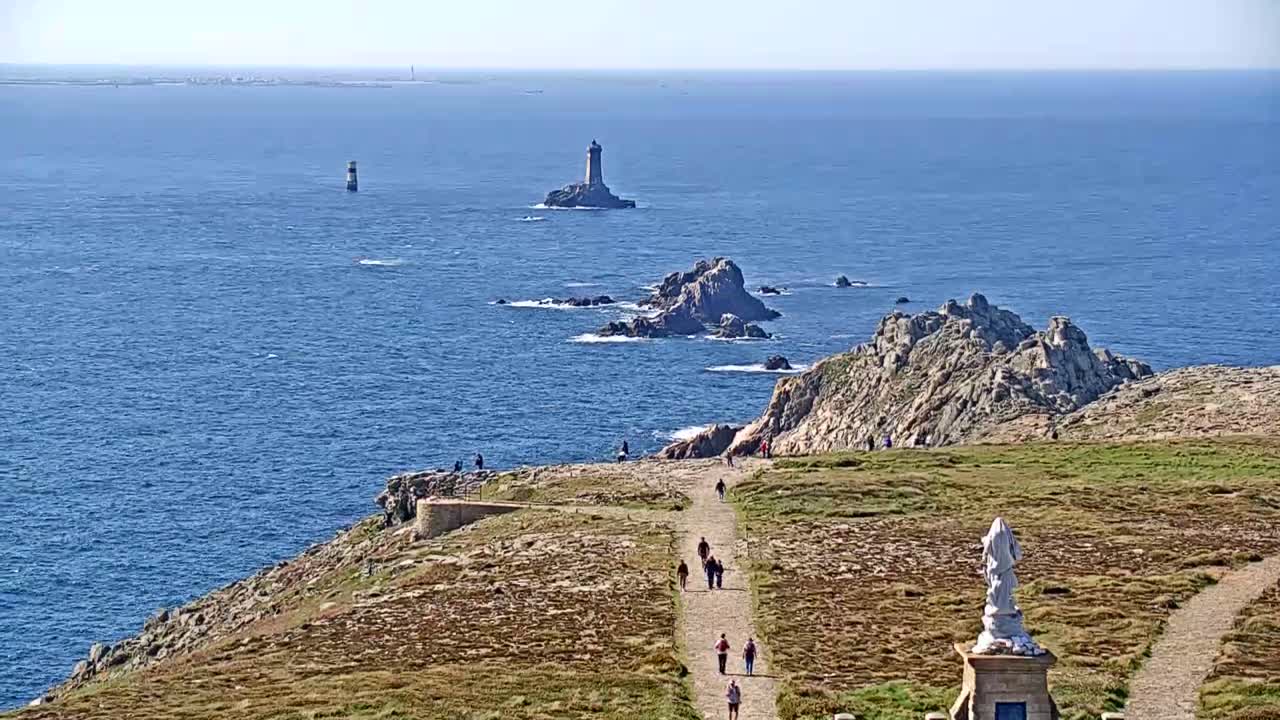 A coastal path winds through scrubland toward a rocky outcrop overlooking a calm blue ocean dotted with several small islands and lighthouses.