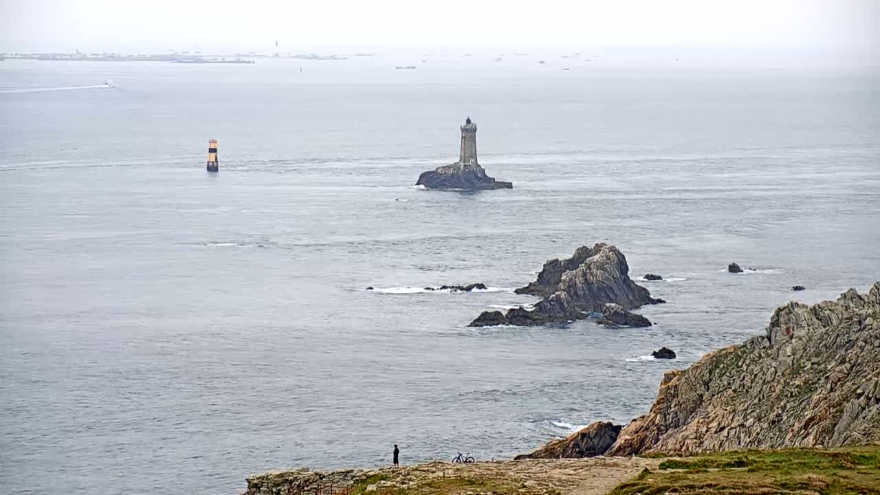 A person stands on a rocky cliff overlooking a calm, gray sea dotted with small islands, one of which holds a lighthouse.