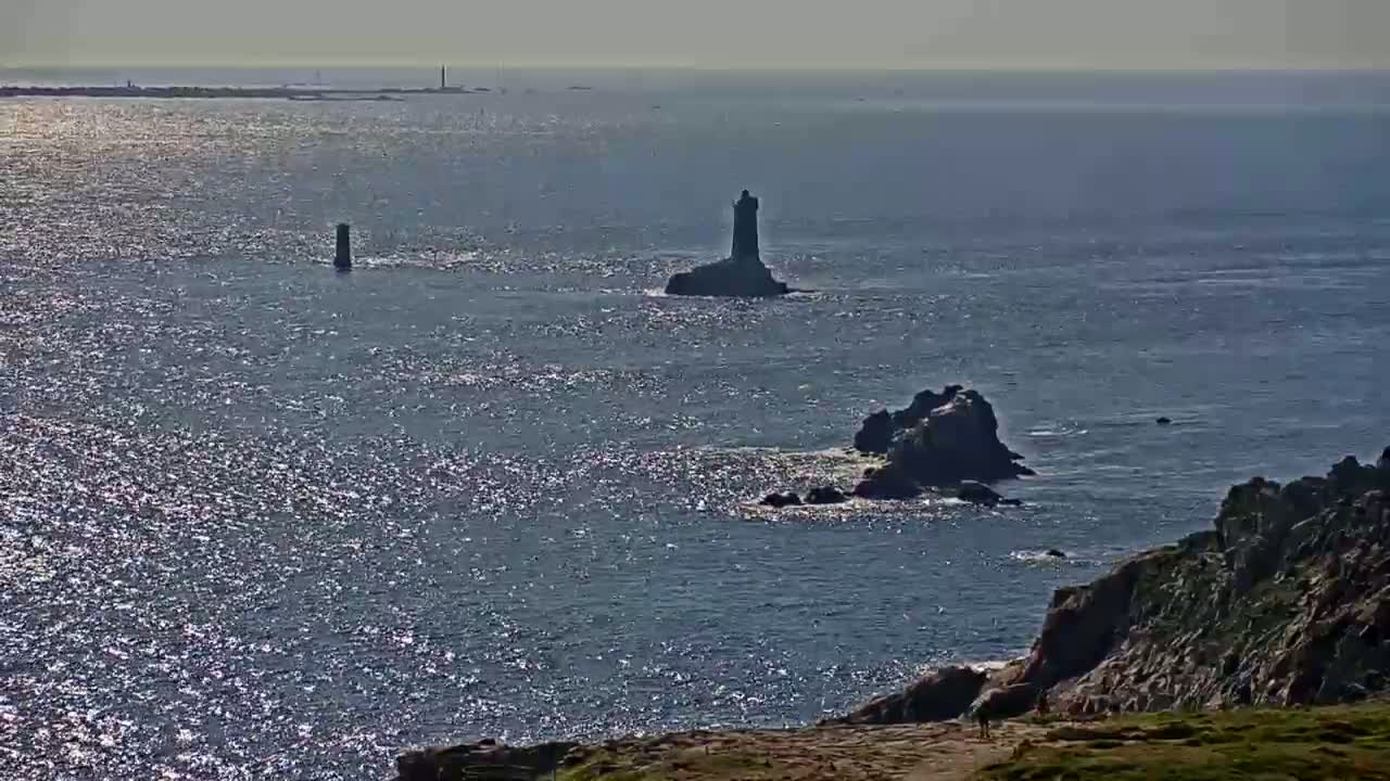 A rocky coastline with a small lighthouse on a distant islet is visible across a sparkling ocean under sunny skies.