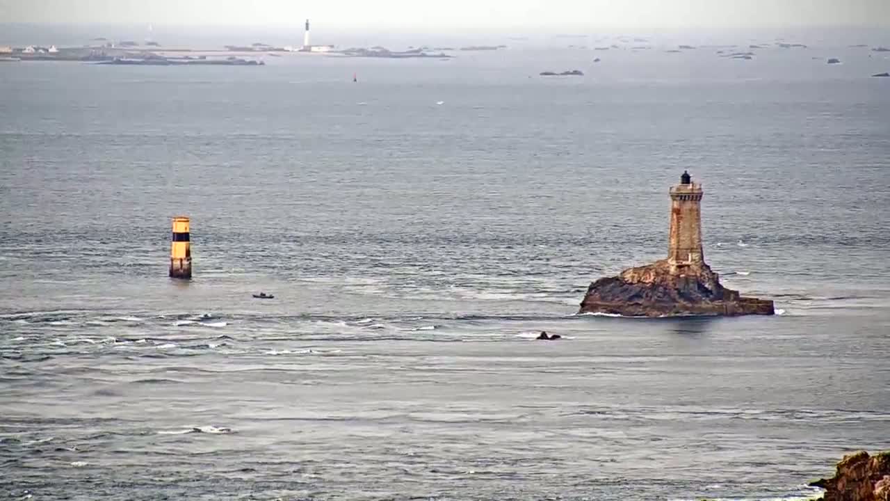 A lighthouse sits on a small rocky island in a choppy sea, with another smaller marker in the distance under an overcast sky.