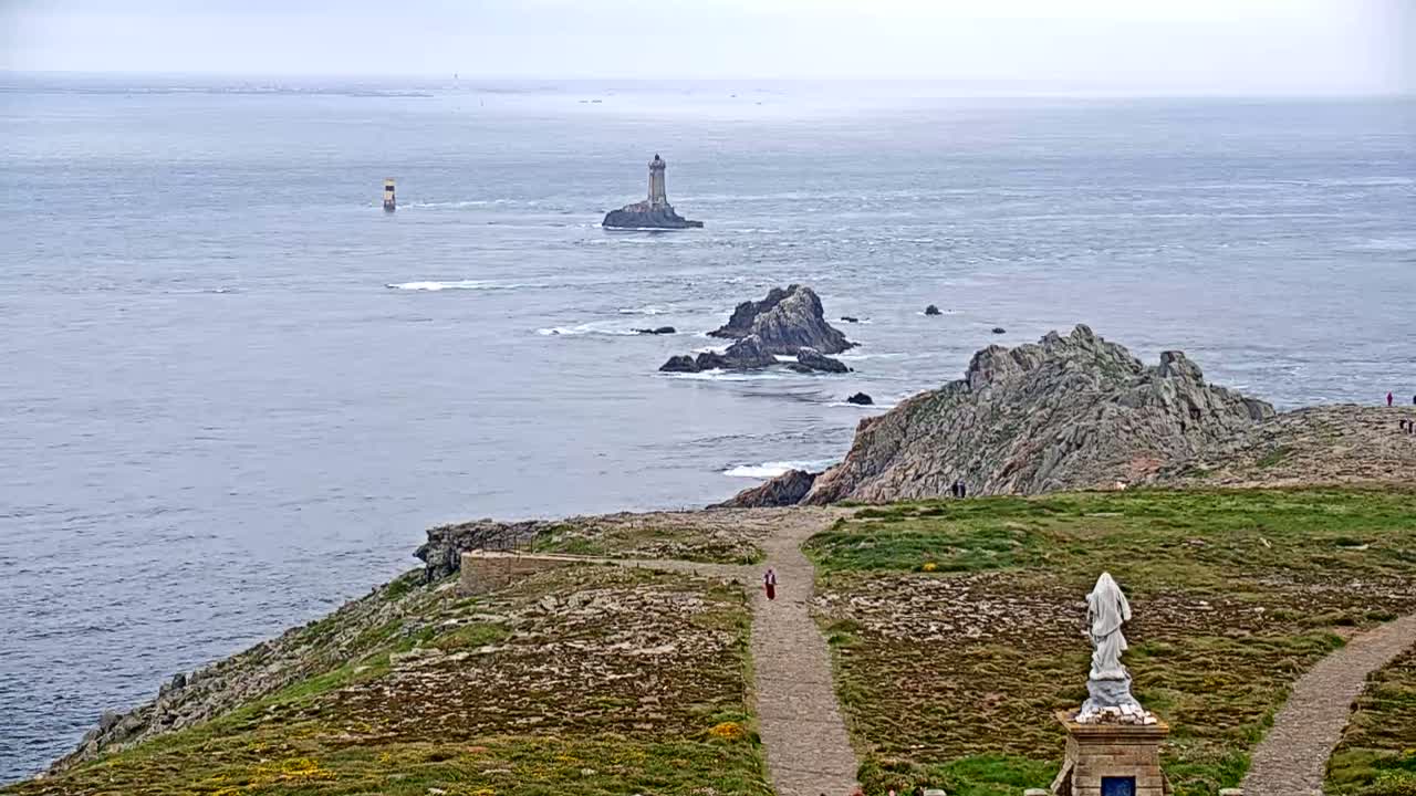 A rocky coastal path overlooks a gray and choppy sea, featuring a distant lighthouse on a small island and a statue on the foreground.