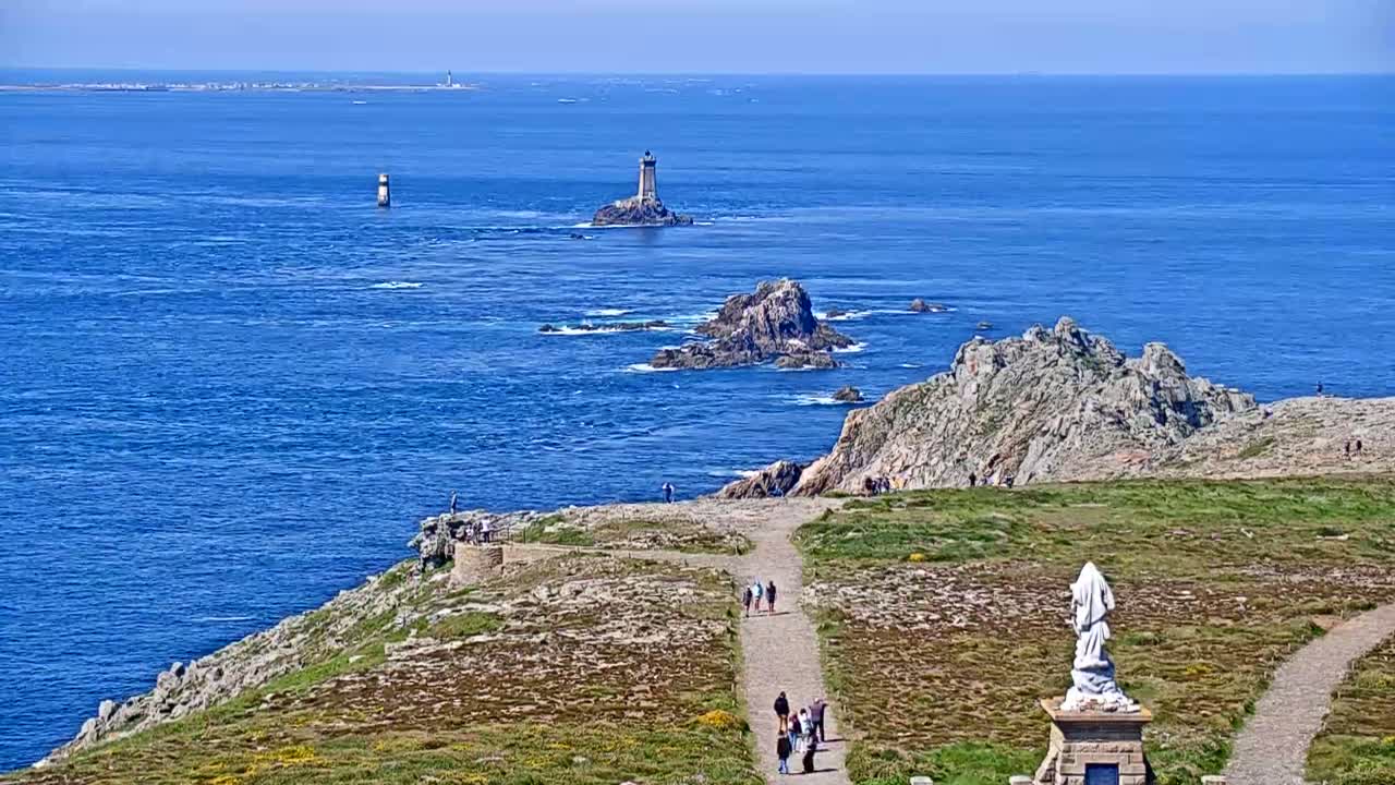 A sunny coastal scene shows a rocky, grassy headland overlooking a vast expanse of blue ocean dotted with islands and lighthouses, with several people walking along a path.