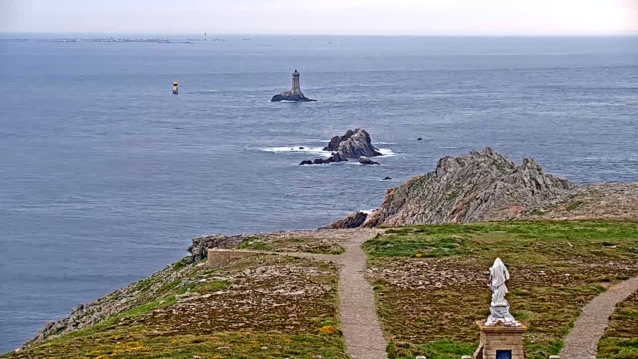 A coastal view shows a statue, a path, rocky terrain, and a lighthouse on an island in the distance on an overcast day.