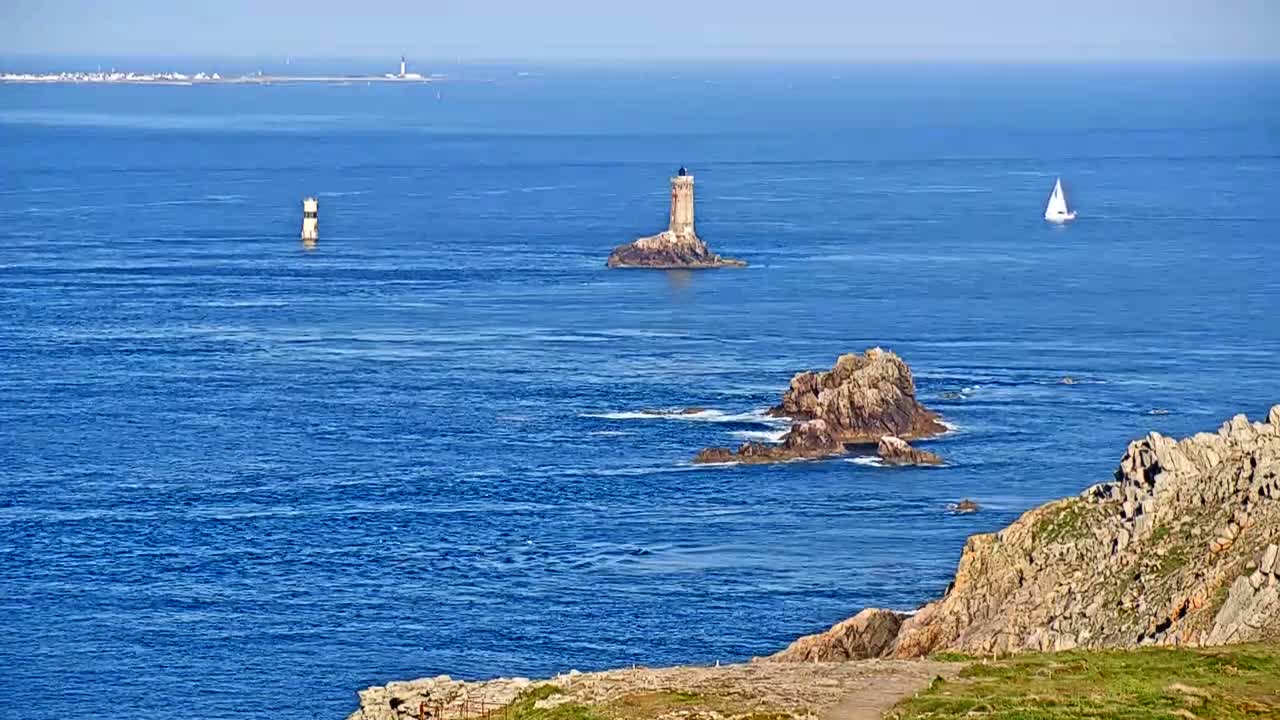 A picturesque coastal scene on a sunny day shows a lighthouse on a small island, another distant lighthouse, a sailboat, and rocky outcrops in a deep blue ocean.