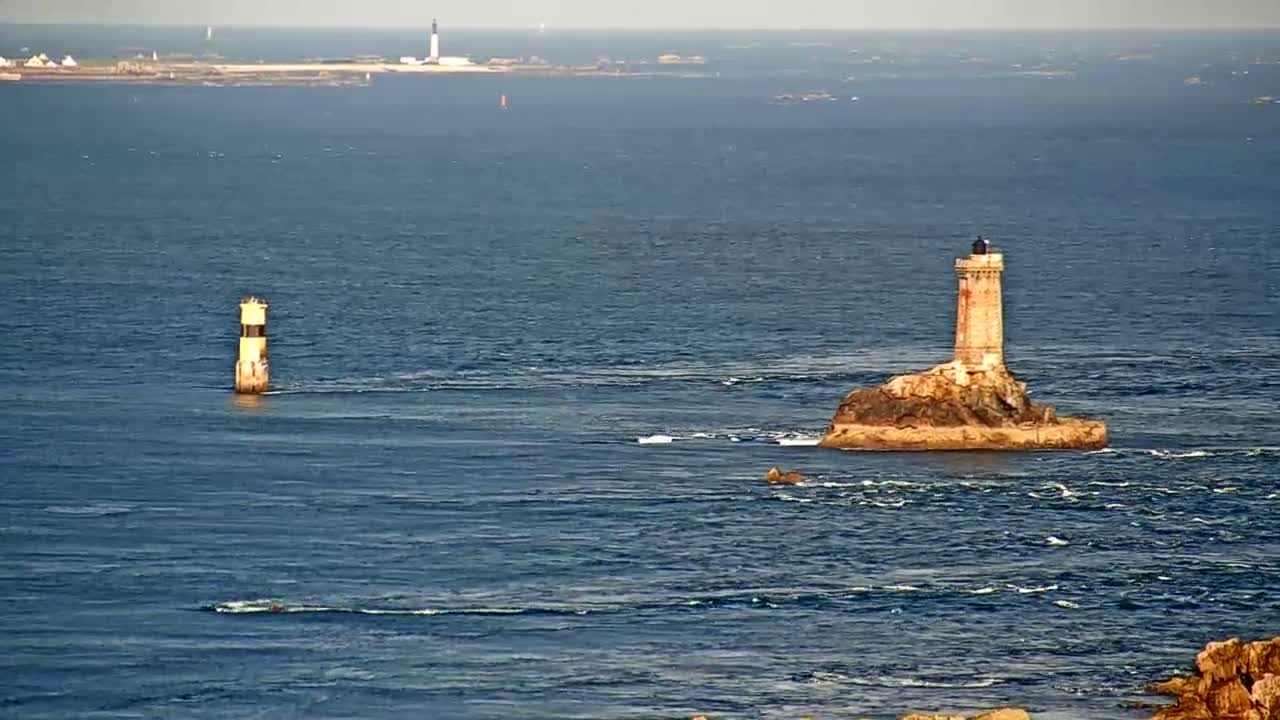 A tall lighthouse sits on a small, rocky island in a calm, blue sea, with a smaller lighthouse visible in the distance.