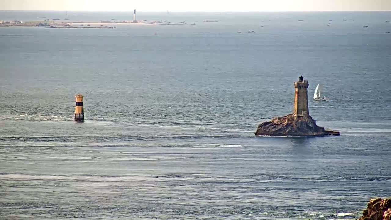 A tall lighthouse sits on a small, rocky island in a calm ocean, with a smaller lighthouse and a sailboat visible in the distance.