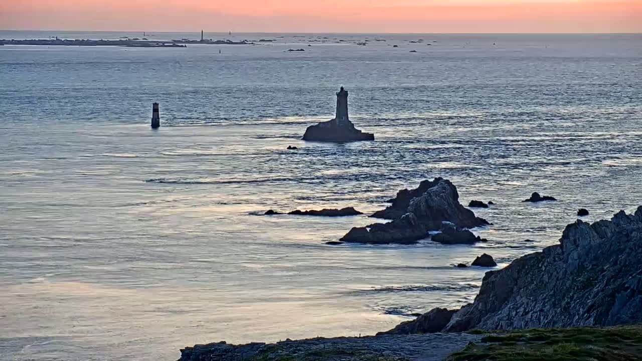 A coastal scene at sunset shows a lighthouse on a small island, rocky outcrops, and calm water.