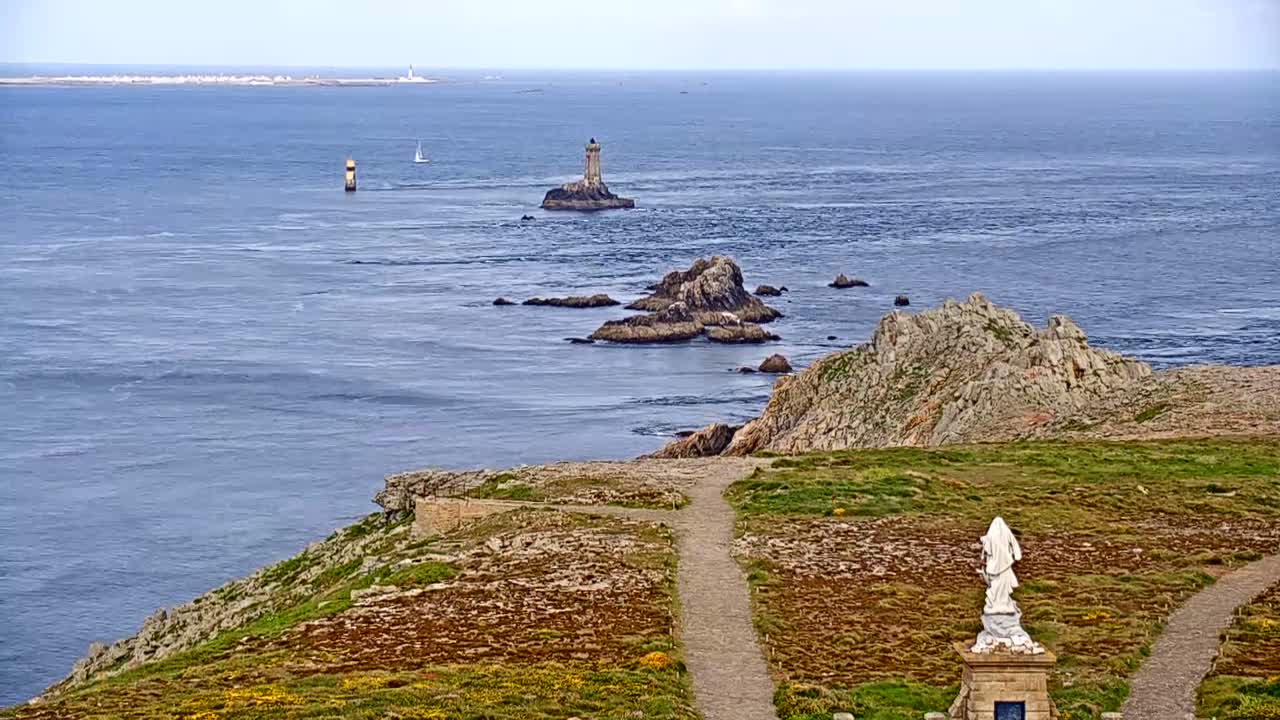 A coastal scene under a mostly clear sky shows a rocky outcrop with a statue, paths, and distant lighthouses and islands on a calm ocean.